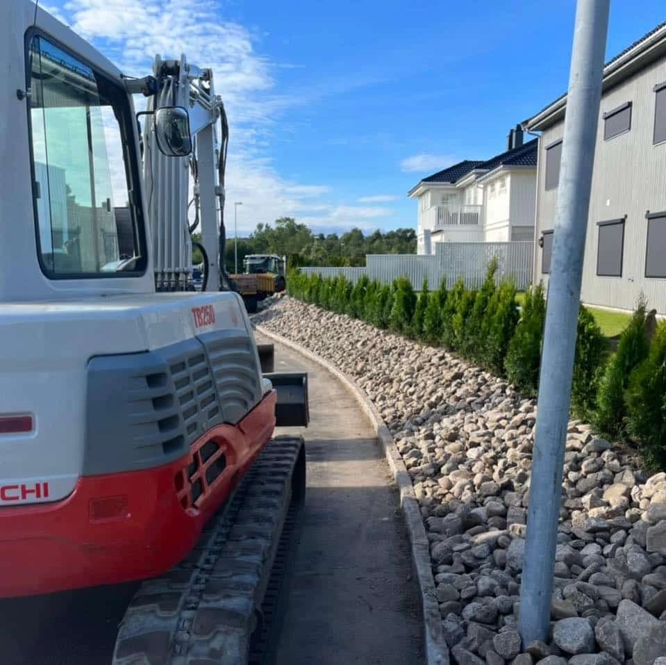 Gravemaskin ved en innkjørsel med steinmasser og hekk langs boliger.