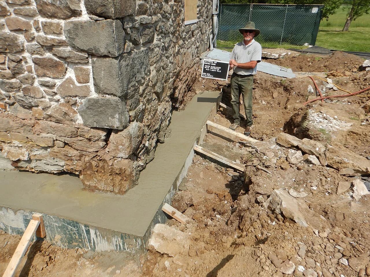En arbeider står ved frilagt murhjørne og reparert betongfundament.
